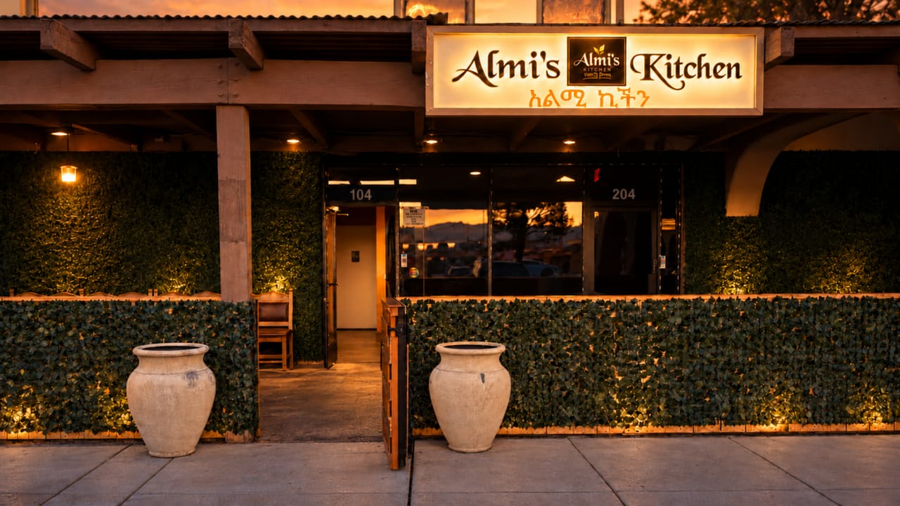The Almi’s Kitchen storefront at sunset: a lit sign in English and Amharic above an open doorway, two cream urns, and a hedge-lined patio under wooden beams.