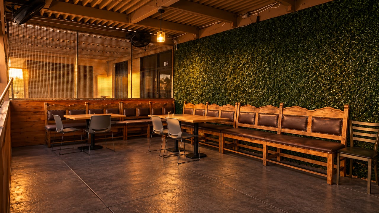 Covered patio with carved wooden benches along a living green wall, light wood tables, and a pendant lamp glowing in the evening sun.